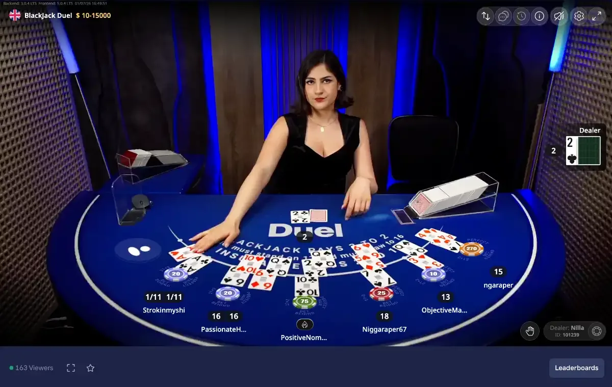 Képernyőkép a Duel Blackjack Live közvetítésről: osztónő a kék Duel asztalnál, a játékosok kiosztott lapjai és zsetonjai, élő nézőszámláló.