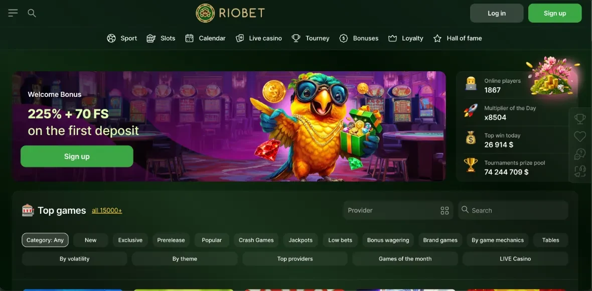 Zrzut ekranu polskiej wersji Riobet Casino (nr 8 w rankingu Kaszinok24): biale logo RIOBET ze ztorym medalionem, gorne menu Sport, Slots, Calendar, Live casino, Tourney, Bonuses, Loyalty i Hall of fame oraz przyciski Log in i Sign up, glowny baner Welcome Bonus "225% + 70 FS on the first deposit" z zielonym przyciskiem Sign up i rysunkowa papuga w okularach trzymajaca zlote monety i prezent na tle lobby kasyna, prawy panel statystyk Online players 1867, Multiplier of the Day x8504, Top win today 26 914 $, Tournaments prize pool 74 244 709 $, sekcja Top games all 15000+ z polem wyszukiwania, wybor kategorii Category: Any, New, Exclusive, Prerelease, Popular, Crash Games, Jackpots, Low bets, Bonus wagering, Brand games, By game mechanics, Tables, By volatility, By theme, Top providers, Games of the month, LIVE Casino (zrzut redakcyjny, 2026).